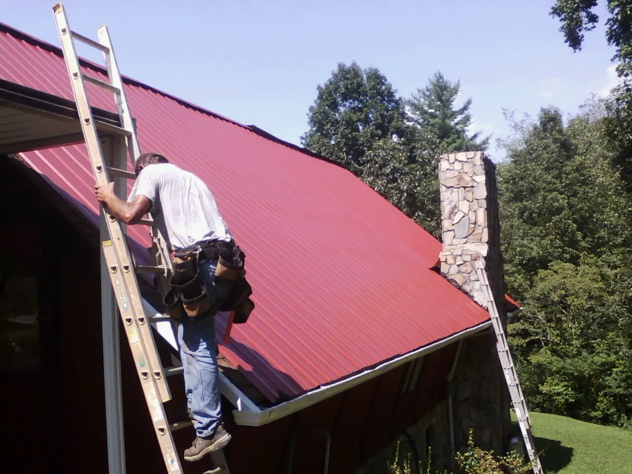 Roofer on a ladder beside a red metal roof and stone chimney — Soffit & Fascia Repair by Roofing Brothers in Laurinburg
